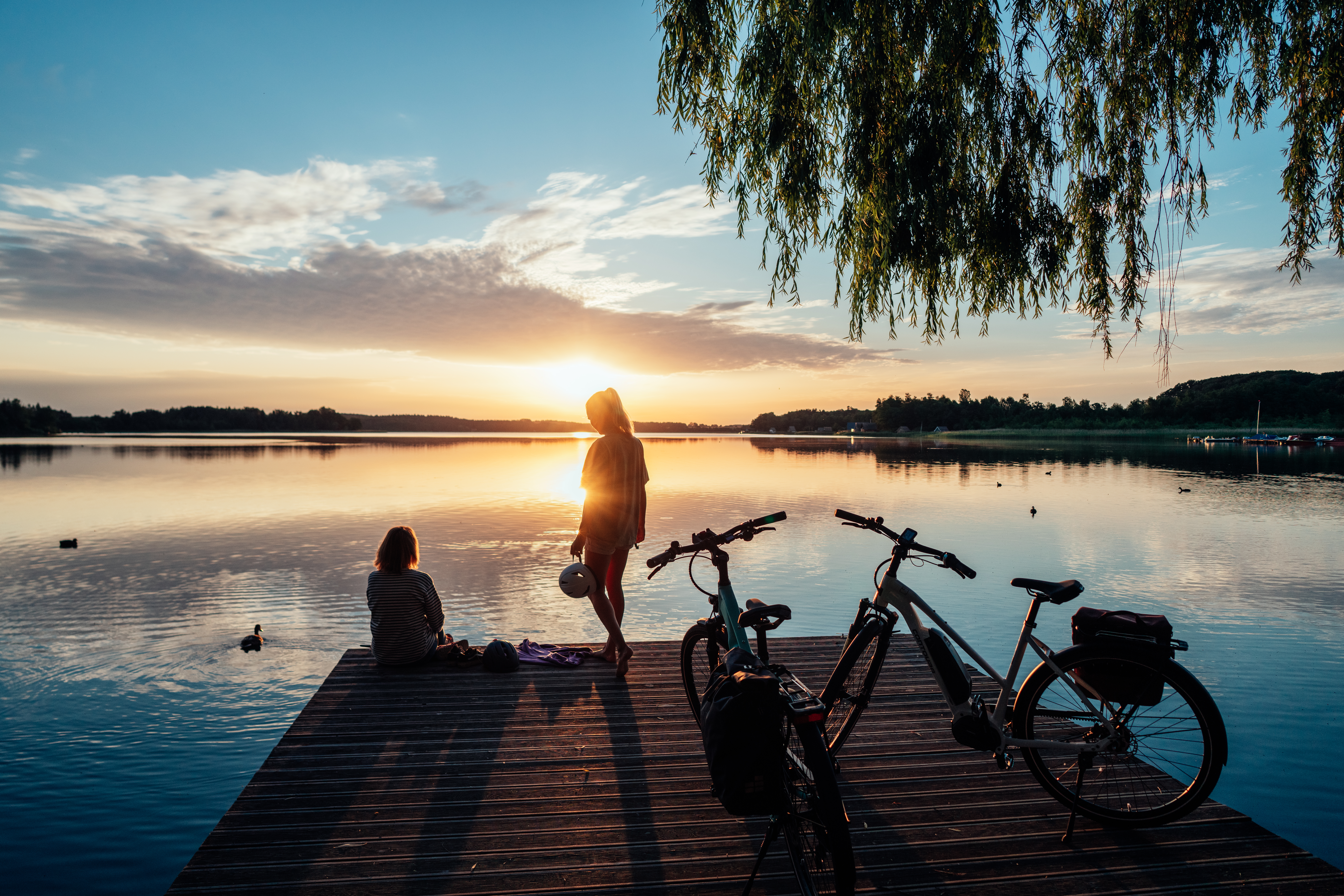 Zwei Frauen befinden sich auf einem Steg der ins Wasser ragt. Eine der Frauen sitzt und blickt aufs Wasser, die andere steht hinter ihr und hält einen Fahrradhelm in der Hand. Langsam geht die Sonne unter und hinter ihnen stehen zwei Fahrräder.